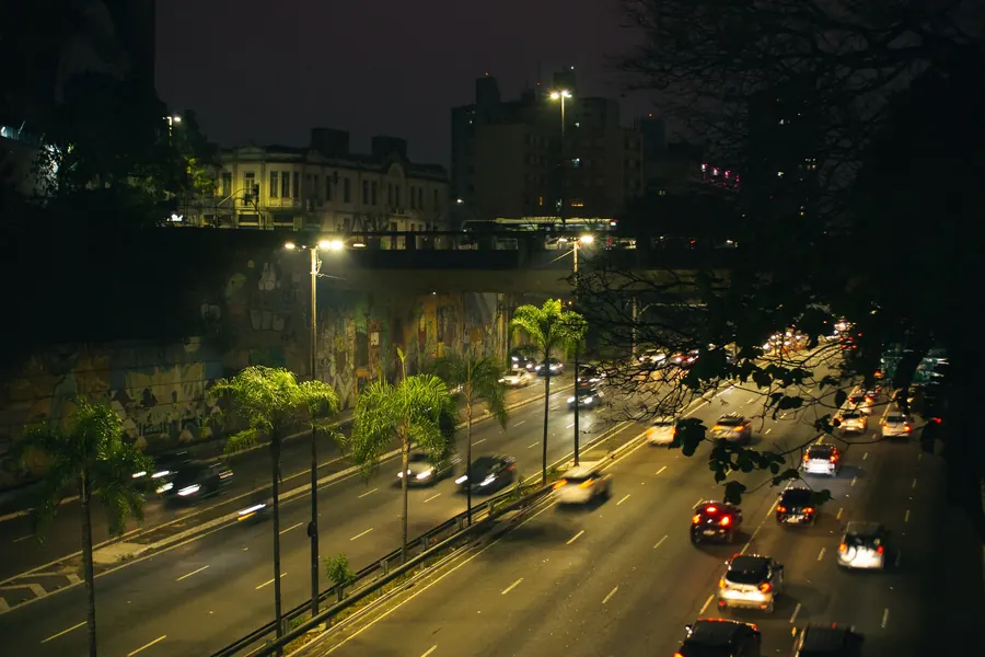 Avenida movimentada à noite, com faróis e palmeiras iluminadas
