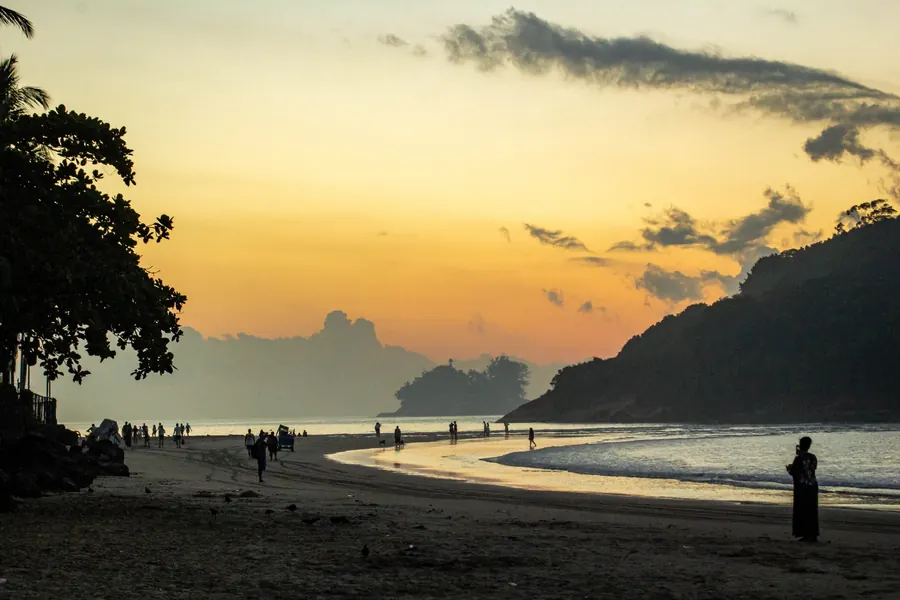 Silhuetas de pessoas na areia ao pôr do sol, com morro e ilha ao fundo